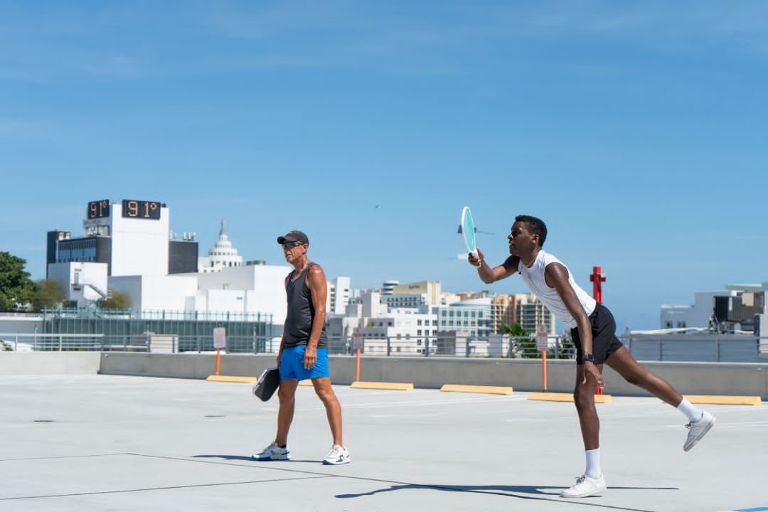 Two queer pickleball players in a rally on a Miami Beach rooftop court, one reaching to return the ball