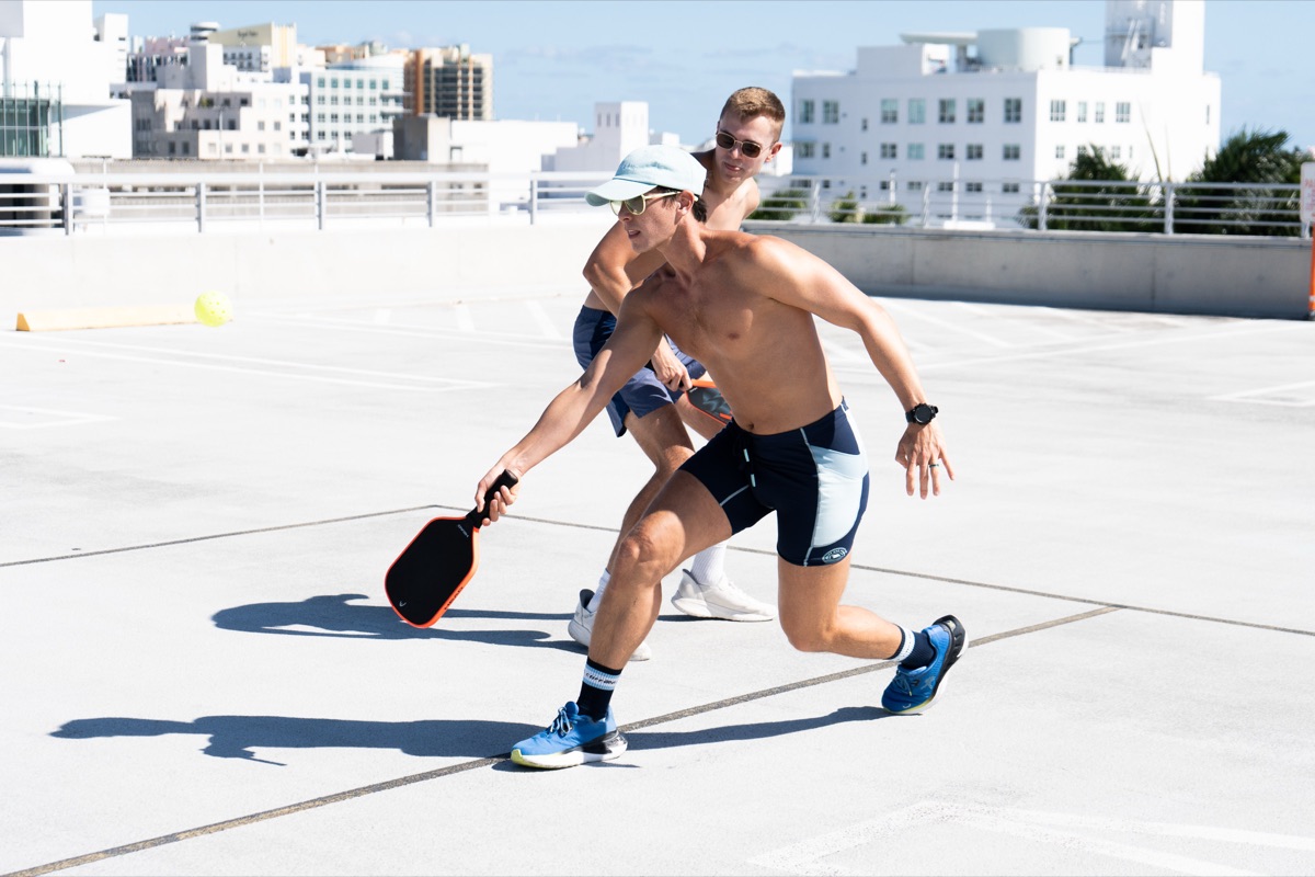 A queer pickleball player lunges for a low shot mid-rally with the Miami skyline behind