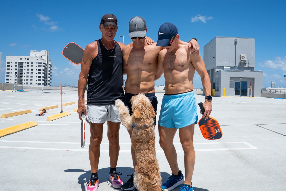 Three LGBTQ+ pickleball players with their paddles and a friendly dog on a Miami Beach rooftop court