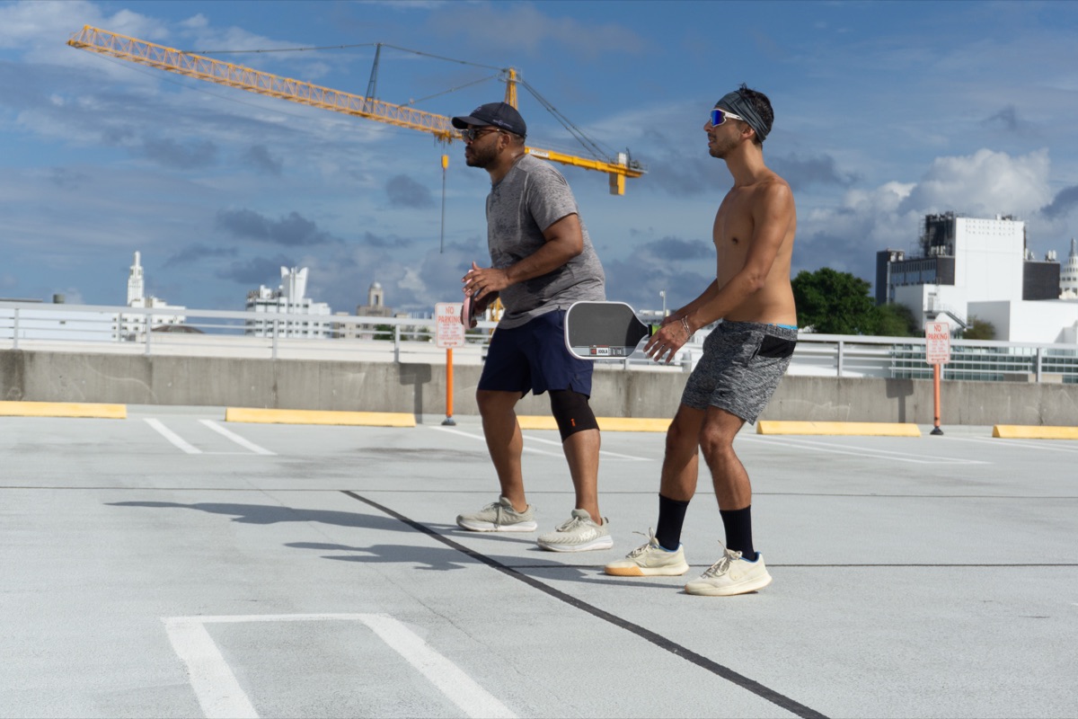 Two gay pickleball players during a game with the Miami skyline and a crane behind them